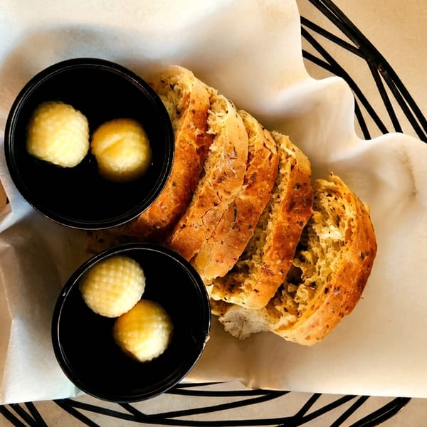 Sliced artisan bread and two small black bowls of butter in a basket lined with parchment paper.