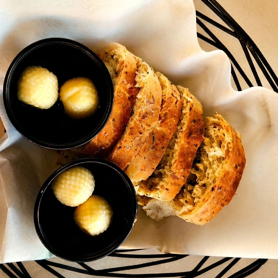 Sliced artisan bread and two small black bowls of butter in a basket lined with parchment paper.