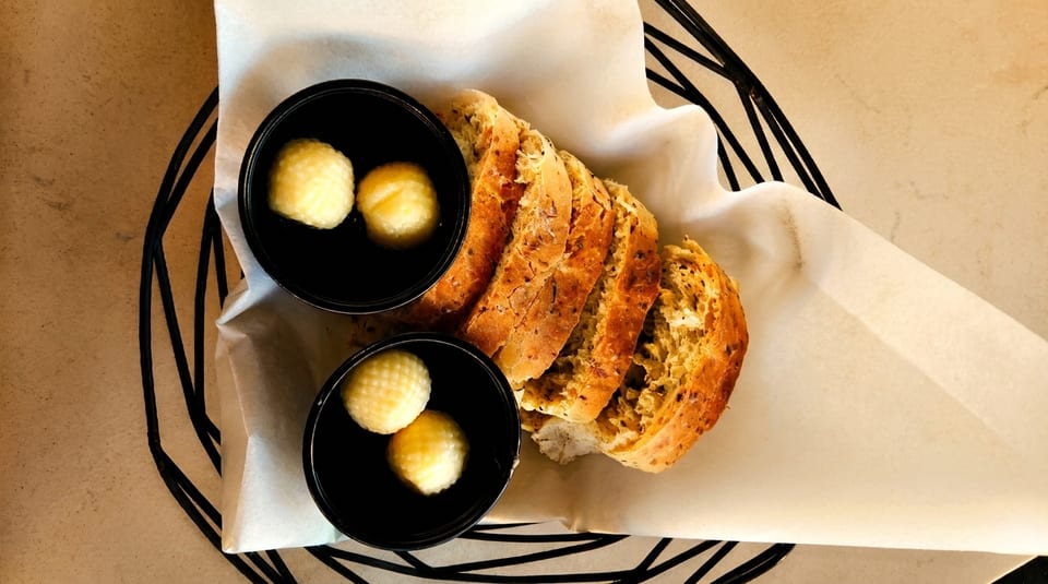 Sliced artisan bread and two small black bowls of butter in a basket lined with parchment paper.