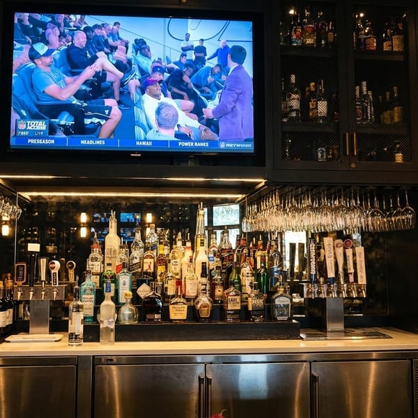 A well-stocked bar with a large TV showing a sports game, liquor bottles, and hanging glassware.