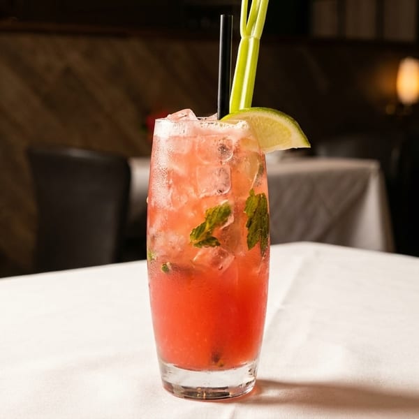 A vibrant red cocktail with ice, mint, lime, and celery on a white tablecloth in a dimly lit restaurant.