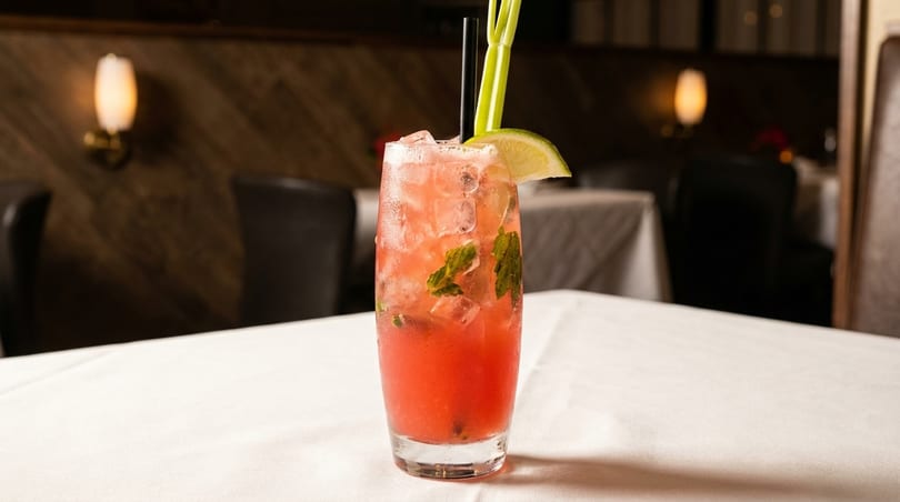 Red cocktail with ice, mint, lime, and celery on a white tablecloth in a dimly lit restaurant.
