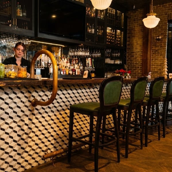 A bartender stands behind a stylish bar with geometric tile, green stools, and brick walls.